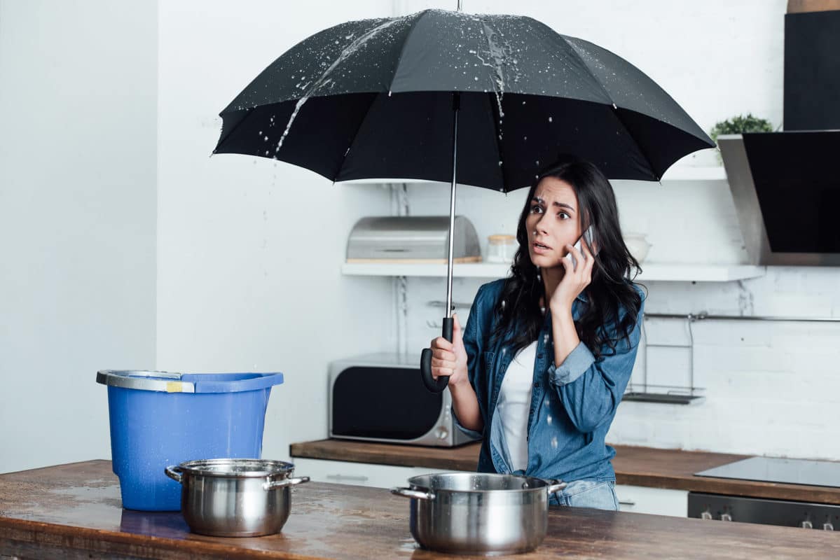 Homeowner on the phone holding an umbrella under a leaking kitchen ceiling